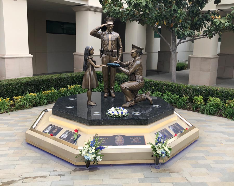 Image of the Buena Park Police departent memorial of fallen officers. Bronze statue of a saluting officer, another officer on one knee presenting a folded flag to a little girl. all cast in bronze.