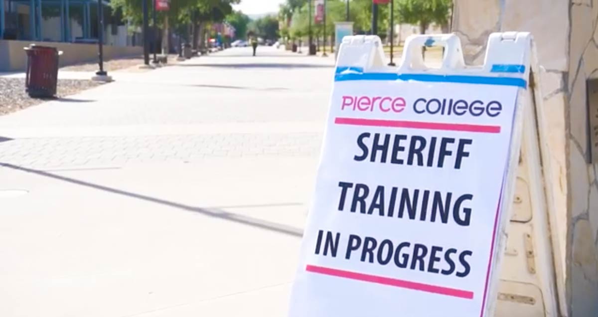 Image of an A-frame sign with big black letters that read "Sheriff training in progress". The sign in the lower left side of the frame. The sign is infront of a college walk path.