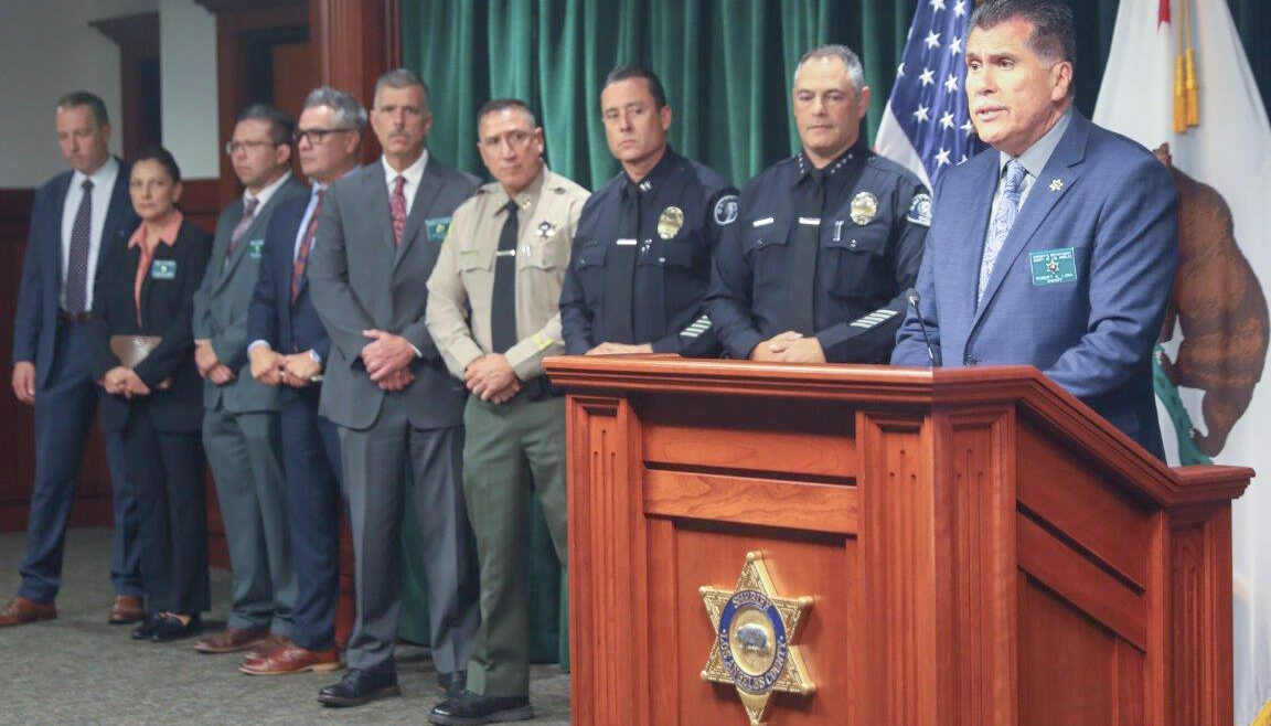 Sheriff Robert luna in a blue suit standing behind a wooden podium. LASD staff as well as LAPD staff stand behind him.