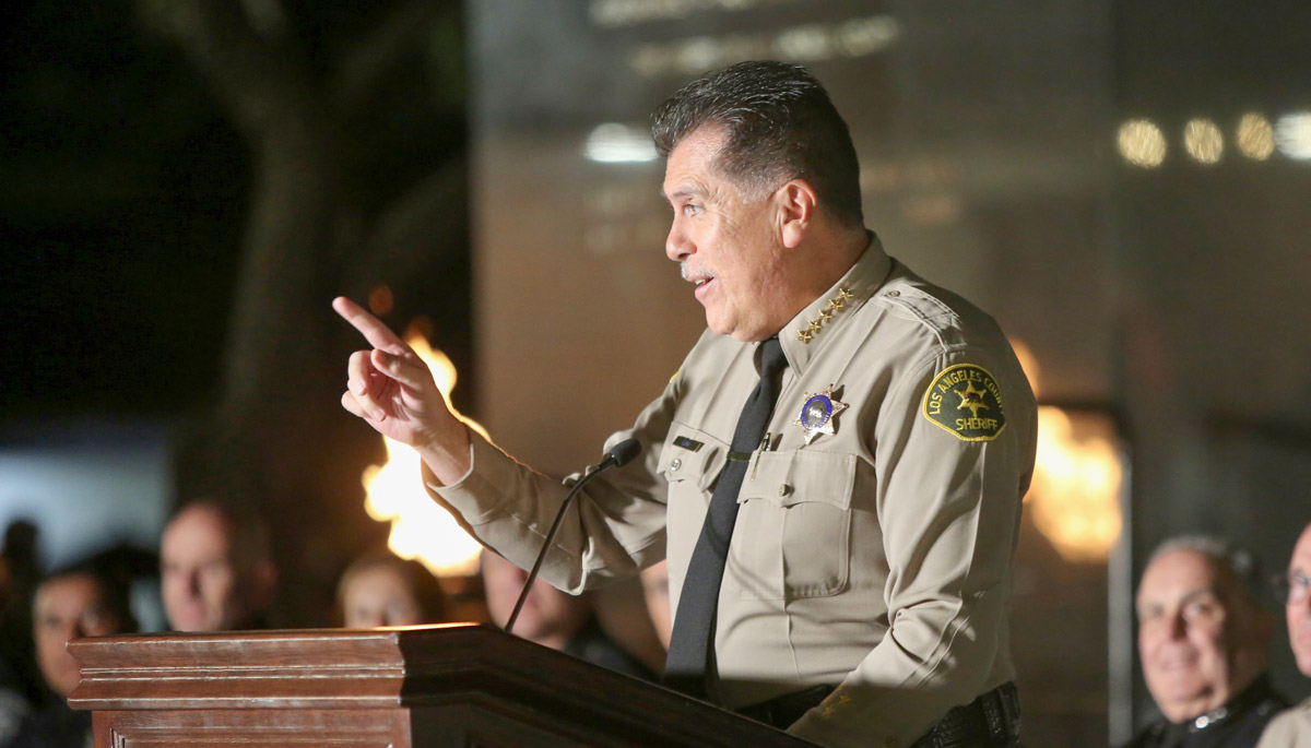 Image of Sheriff Robert Luna, standing at a podium speaking to a crowd that is out of frame, He is pointing one finger outward towards the audience. He is wearing a tan uniform shirt with a black tie and a badge over his left side. 