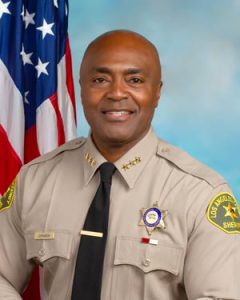 Assistant Sheriff Johnson is wearing a tan long sleeve shirt with a black tie, badge on left side of shirt, sitting in front of a blue background. There is a hanging American flag in the background behind him on his right side.