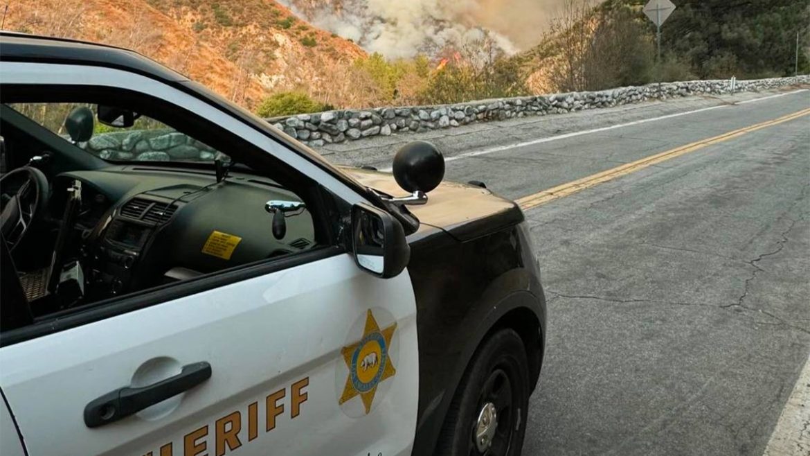 Image of a mountain road with at Sheriff's Black and White vehicle, parked on the road on the lower left of image. The trees beyond the road are on fire and smoke is billowing up across the hills.