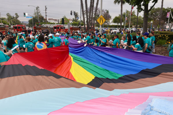 Parade of people walking a pride flag the size of the street. The street is filled with the flag up to the top of the image where people are holding it.