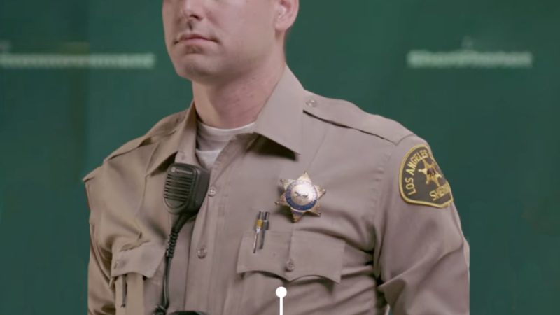 Class A Patrol Uniform: deputy with sandy blond hair is posing. he is wearing a tan wool longsleeve button down shirt. there is a gold LA County Sheriff's badge over his left pocket. There is a black rectangluar Body Camera clipped to his shirt in the middle. He has a walkie Talkie clipped to his lapel.